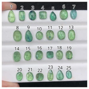 May include: A collection of twenty-five green gemstones in various shapes, including oval, pear, and square cuts. The gems are arranged in rows, showcasing their different facets and shades of green, from light to deep teal.