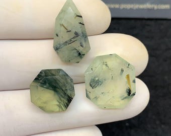 Könnte beinhalten: Drei facettierte Prehnit-Edelsteine in verschiedenen Formen. Die Edelsteine sind hellgrün mit dunklen Einschlüssen. Die Edelsteine werden auf einer weißen Oberfläche präsentiert. Der Text "POPE JEWELRY" ist im Hintergrund sichtbar.