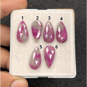 May include: Six faceted pear-shaped gemstones arranged on a white display tray. Each stone features a translucent base with vibrant pink inclusions and distinct geometric facets. The stones are numbered one through six in black text above each piece.