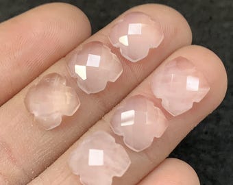 Puede incluir: Seis cuentas de gemas facetadas en forma de trébol de color rosa pálido. Cada cuenta tiene un corte geométrico que refleja la luz. Las gemas se sostienen en una mano y tienen una calidad translúcida.
