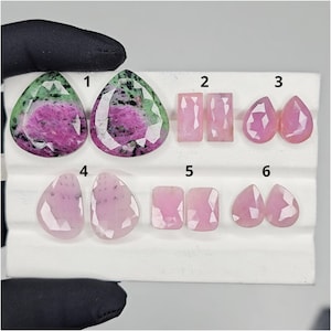 May include: A collection of six faceted gemstones in various shapes. Two pear-shaped stones with green and pink patterns, and four pairs of pink gemstones in rectangular, teardrop, and irregular shapes. The gems are displayed on a white surface.