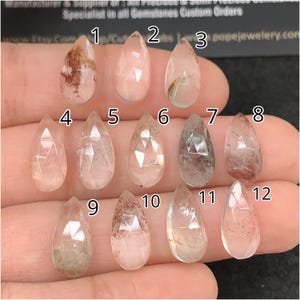 May include: A collection of twelve teardrop-shaped gemstones, each with unique internal inclusions and facets. The gems vary in color, from clear to pale pink and brown, with visible patterns. The gemstones are displayed against a neutral background.