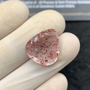 May include: A clear, triangular gemstone with red inclusions. The gem is held in a gloved hand, showcasing its clarity and the intricate patterns within. The image is well-lit, highlighting the stone's natural beauty. The background is blurred, focusing attention on the gem.