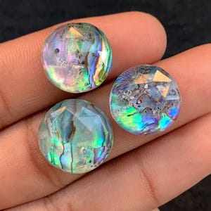 May include: Three iridescent abalone shell cabochons with faceted surfaces. The cabochons display blue, green, and gold colours, and are round. The gems are held in a hand against a dark background.
