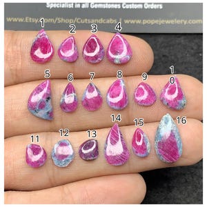 May include: A collection of sixteen polished ruby and zoisite cabochons arranged in rows, each numbered from 1 to 16. The stones feature vibrant magenta-pink ruby inclusions set within a mottled grey and blue-green zoisite matrix.