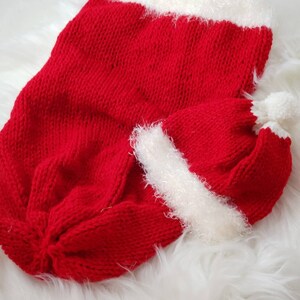 May include: Red knitted Santa hat and matching outfit for a newborn baby. The hat has a white pom-pom on top and the outfit has a white fuzzy trim.