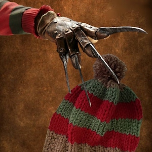 May include: A red and green striped knit beanie with a brown pom pom on top. The beanie is being held by a clawed hand with metal blades.
