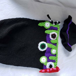 May include: A black and green knitted baby sleeper with a purple and green eye design and a matching black hat with a purple trim.