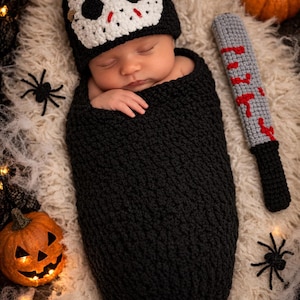 Puede incluir: Un beb&eacute; reci&eacute;n nacido envuelto en un capullo negro de ganchillo, con un gorro de m&aacute;scara de hockey blanco y negro. Un accesorio de arma gris y rojo de ganchillo y dos calabazas completan el tema de Halloween.