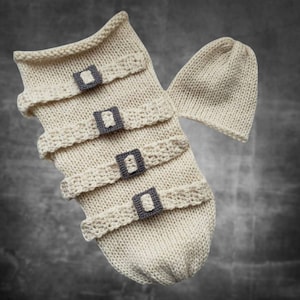 May include: A knitted baby sleeping bag with a matching hat. The sleeping bag is cream colored and has four black buckles. The hat is also cream colored.