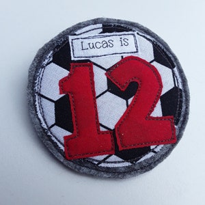 May include: A grey felt circle with a black and white football design. The number 12 is in red felt on top of the football design. A white rectangle with the text "Lucas is" is on top of the football design.