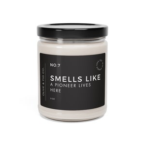 May include: A glass jar candle with a black label that reads "Olive & Fig Co. No. 7 Smells Like A Pioneer Lives Here 9 oz". The candle is 100% soy and hand-poured.