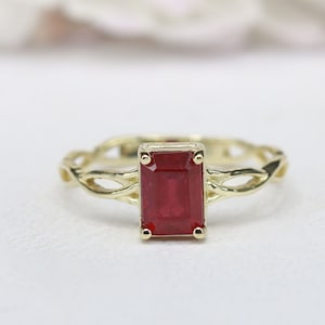 Könnte beinhalten: Ein goldener Ring mit einem rechteckigen, tiefroten Edelstein. Das Band des Rings hat ein aufwendiges, gewebtes Design. Der Edelstein ist in einer goldenen Lünette gefasst. Der Ring wird auf einer weißen Oberfläche präsentiert.