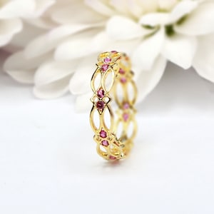 Könnte beinhalten: Ein goldener Ring mit einem filigranen, durchbrochenen Design und rosafarbenen Edelsteinen. Der Ring ist vor einem weißen Hintergrund platziert.