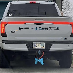 以下が含まれることがあります： 白いフォードF-150ライトニングトラックの後部ビュー。トラックは赤いライトバー、F-150のロゴ、BUZZ LTGと書かれたナンバープレートを備えています。青い充電ポートカバーがヒッチからぶら下がっています。