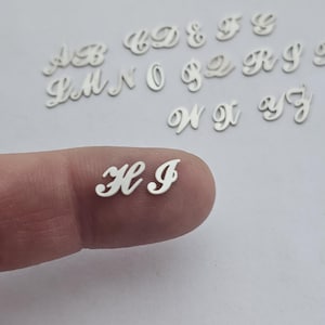 May include: A close-up of a silver letter H and I, in a cursive font, on a finger. The letters are small and delicate, and they are perfect for adding a personal touch to jewelry or crafts.