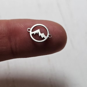 May include: Silver charm with a mountain silhouette design inside a circle. The charm has two jump rings for attaching to a bracelet or necklace.