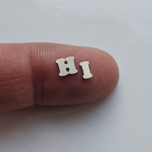 Op de afbeelding: Close-up van twee kleine, zilveren metalen letters die "HI" spellen op een vinger.