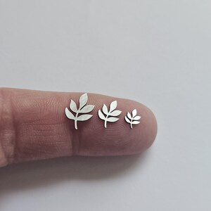 May include: Three silver-colored metal leaf-shaped charms of varying sizes arranged in a row. Each charm features a central stem with symmetrical, pointed leaves extending from the sides, showcasing a flat, smooth, and reflective metallic surface.