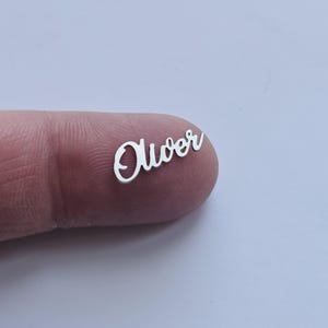 Op de afbeelding: Een klein, zilveren, cursief naamplaatje met het woord "Oliver" uitgesneden.