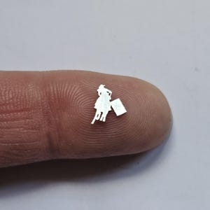 May include: A tiny, silver-colored metal cutout of a person holding a shopping bag. The figure is wearing a dress and has a hat. The cutout is resting on a fingertip, with a blurred background.