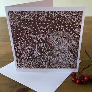 Peut inclure: Une carte de vœux marron avec un dessin au trait blanc d'un lapin dans un champ d'herbe haute et de fleurs. L'arrière-plan est de couleur marron foncé avec des points blancs.