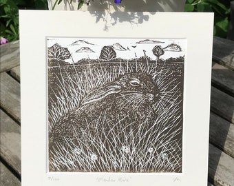 May include: A black and white linocut print of a meadow hare lying in tall grass. The print is titled "Meadow Hare" and numbered 9/100.