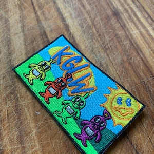 Peut inclure: Écusson brodé avec quatre personnages extraterrestres colorés et un soleil souriant sur un fond bleu et vert. L'écusson porte le texte "KGW" en orange et bleu. L'écusson est rectangulaire avec une bordure noire.