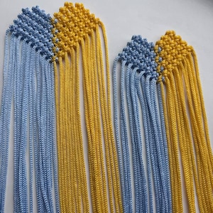 May include: Two macrame pieces featuring a diamond-shaped knot pattern at the top, transitioning into long, loose hanging cords. The design incorporates alternating sections of light blue and bright yellow cord, creating a vibrant, dual-colored effect.