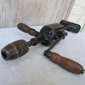 May include: An antique hand drill with a black metal body and wooden handles. The drill has a chuck at the end for holding drill bits. The wooden handles are a dark brown color. The drill is designed to be operated by hand.