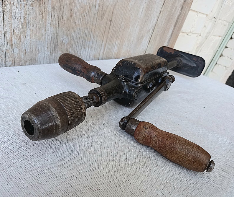 May include: An antique hand drill with a black metal body and wooden handles. The drill has a chuck at the end for holding drill bits. The wooden handles are a dark brown color. The drill is designed to be operated by hand.