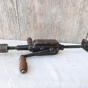 May include: An antique hand drill, featuring a black metal body with a wooden handle and a chuck for holding drill bits. The drill has a crank for manual operation and a small, flat plate on the end.