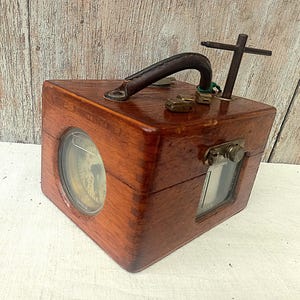 Op de afbeelding: Een vintage houten kist met een leren handvat en een kompasachtig wijzerplaat. De kist heeft een rechthoekige vorm met een klein venster en metalen hardware. Het hout heeft een warme, roodbruine tint.