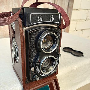 Op de afbeelding: Een vintage twin-lens reflexcamera met een bruine leren hoes en riem. De camera heeft twee lenzen, een zwarte behuizing en de tekst "HAIOU SHANGHAI CHINA" is zichtbaar. De camera heeft ook de Chinese karakters "海鸥".