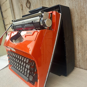 May include: A vintage orange Tippa typewriter with a black keyboard and silver detailing. The typewriter is in its black carrying case. The Adler brand is visible on the roller. The keys and roller are visible.