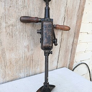 May include: An antique hand drill with a dark metal body and wooden handles. The drill has a chuck at the top and a foot at the bottom for stability. The wooden handles are a warm brown color, contrasting with the dark metal.