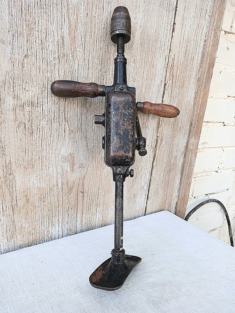 May include: An antique hand drill with a dark metal body and wooden handles. The drill has a chuck at the top and a foot at the bottom for stability. The wooden handles are a warm brown color, contrasting with the dark metal.