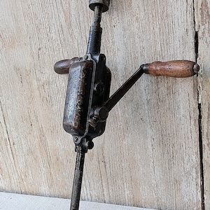 May include: An antique hand drill, featuring a dark metal body with wooden handles. The drill has a crank handle and a chuck for holding drill bits. The base is a flat, rectangular shape. The tool shows signs of age and use.