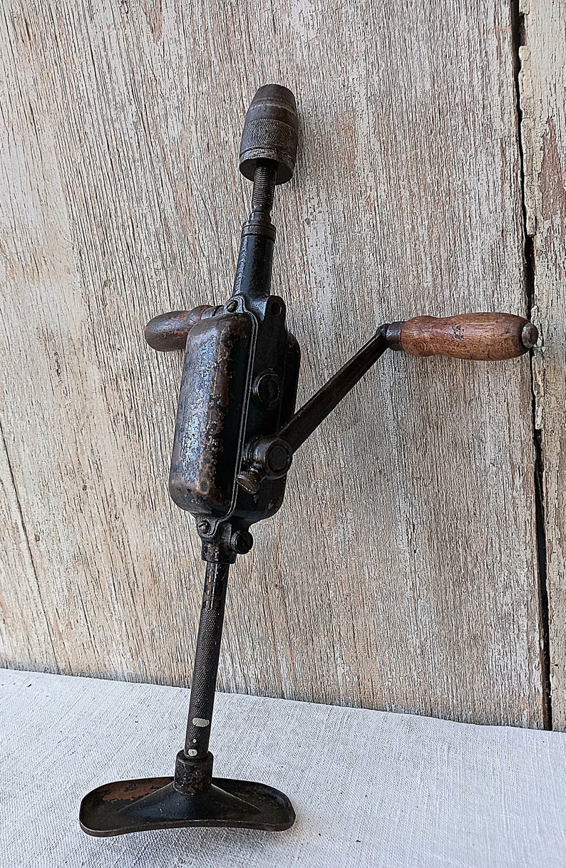 May include: An antique hand drill, featuring a dark metal body with wooden handles. The drill has a crank handle and a chuck for holding drill bits. The base is a flat, rectangular shape. The tool shows signs of age and use.