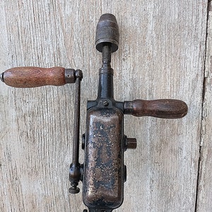 May include: An antique hand drill with a dark metal body and wooden handles. The drill has a chuck at the top and a crank handle on the side. The wood handles are a light brown color.