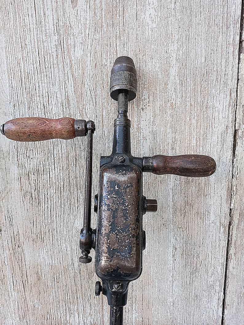 May include: An antique hand drill with a dark metal body and wooden handles. The drill has a chuck at the top and a crank handle on the side. The wood handles are a light brown color.