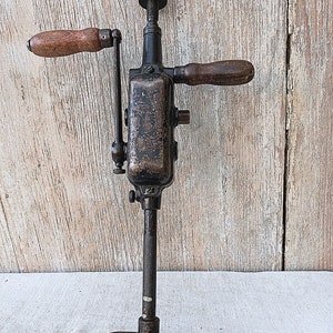May include: An antique hand drill with a dark metal body and wooden handles. The drill has a crank handle on one side and a wooden handle on the other. The base is a flat, curved shape. The drill is designed for manual operation.