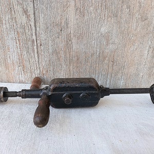 May include: An antique hand drill with a black metal body, wooden handles, and a chuck. The drill has a rusty appearance, suggesting age and use. The tool is designed for manual operation and is likely used for woodworking or similar tasks.