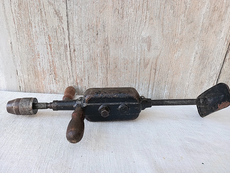 May include: An antique hand drill with a black metal body, wooden handles, and a chuck. The drill has a rusty appearance, suggesting age and use. The tool is designed for manual operation and is likely used for woodworking or similar tasks.