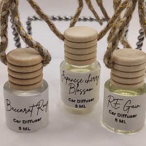 May include: Three car diffusers with wooden lids and labels. The labels read "Baccarat Rouge", "Japanese Cherry Blossom", and "RE Gain". Each bottle contains 5 ML of liquid. The diffusers are suspended by a rope with gold accents.