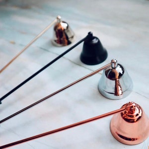 May include: Candle snuffers in silver, black, and rose gold. Each snuffer has a bell-shaped head and a long handle. The candle accessories are arranged on a light-colored surface, offering a modern and elegant aesthetic for home decor.