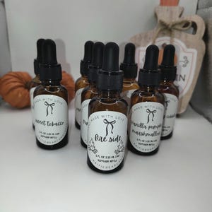 May include: A collection of brown glass bottles with black droppers, labeled with text including "Sweet Tobacco", "Fire Side", and "Vanilla Pumpkin Marshmallow". Each bottle has a white label with a bow design. The bottles are diffuser refills.