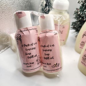 May include: Two pink foaming soap refill sets, each in a clear plastic wrapper. The bottles have white tops and feature the text "Ginger Snap Scent" and "Cinnamon Stick Scent". Other bottles are visible in the background.