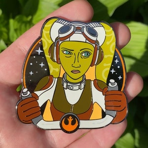 May include: A gold-colored enamel pin featuring a cartoon portrait of a female character with green skin, wearing a white helmet and goggles, and a brown flight suit. The character is holding a steering wheel with a Rebel Alliance symbol in the center. The background is a black and white space scene with stars.