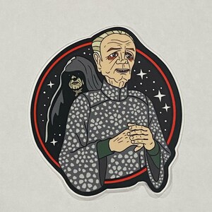 May include: A sticker featuring a cartoon illustration of a man with white hair and a grey patterned robe. He is standing in front of a black background with stars and a red circle. The man is looking to the right and has his hands clasped in front of him. The man is wearing a green undershirt. The sticker is a circular shape with a white border.